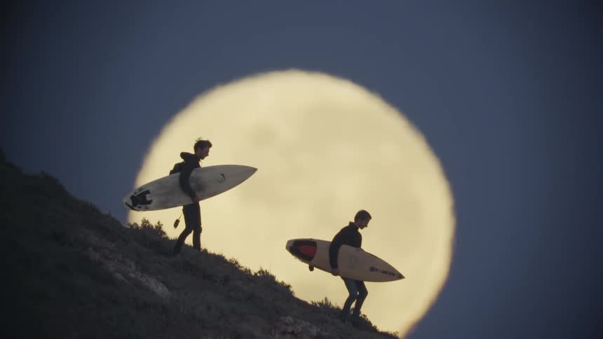 Two men are walking down a steep hill carrying surfboards as the rising full moon illuminates the sky behind them
