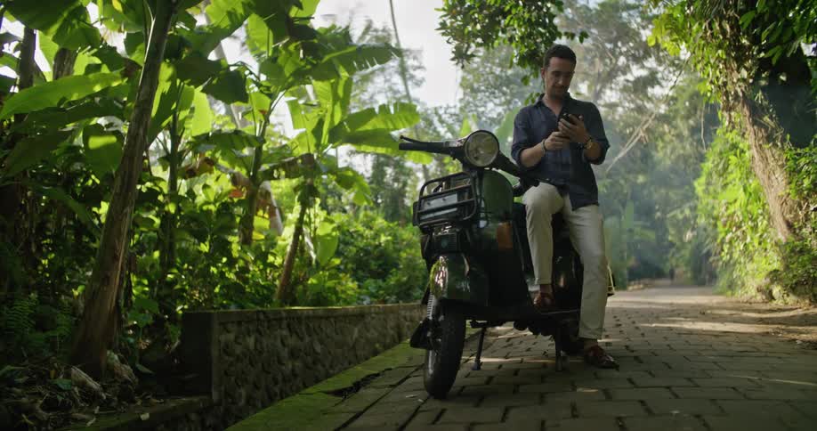 A man is sitting on his motorbike on a cobblestone road and looking at his cell phone and looking up at the trees