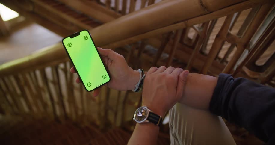 Man with dark blue shirt and brown pants and wristwatch sits on stairs next to wood railings using smartphone