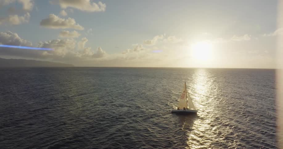 Aerial view of a small sailboat sailing on the calm surface of the ocean at sunset