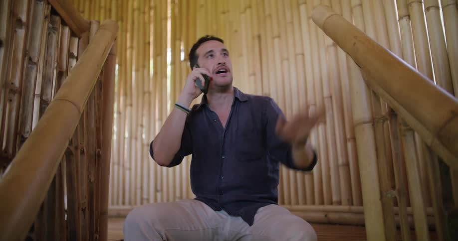 Man sits at top of steps in area constructed of bamboo and talks on smartphone