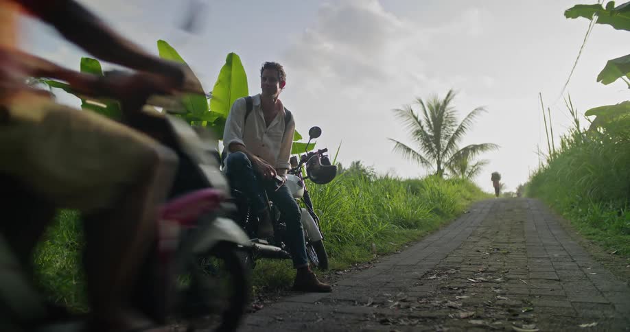 A man sitting on a motorcycle parked beside a narrow rural road raises his camera to take a photograph