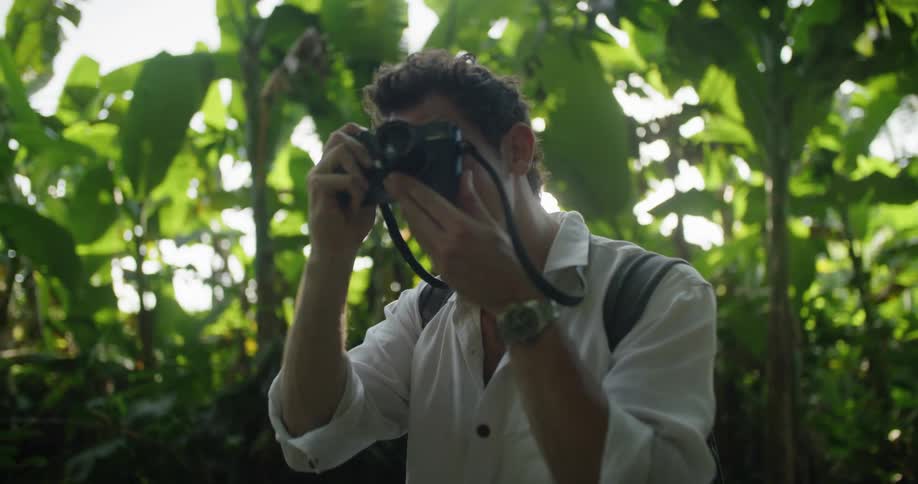 Man with white shirt black backpack and silver watch takes photographs with camera in jungle