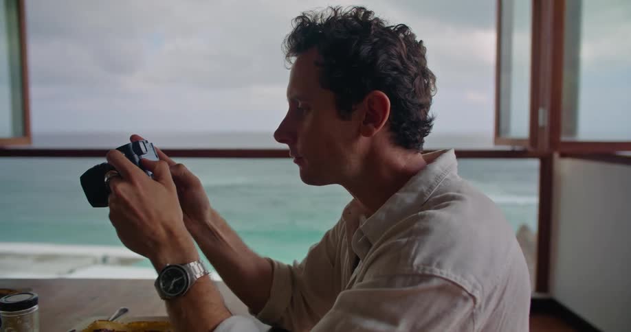 Man chews and examines camera while sitting at dining table near open window with view of coast