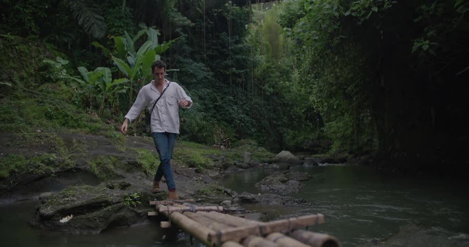Man fords river using bamboos lashed together as large bridge towers overhead
