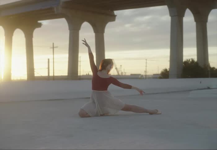 Young female ballet performer dances on the roof of a building near a bridge at sunset