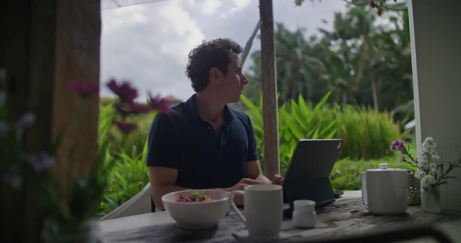 A man sitting at a patio counter drinking coffee and typing on a laptop with lush green tropical plants behind him