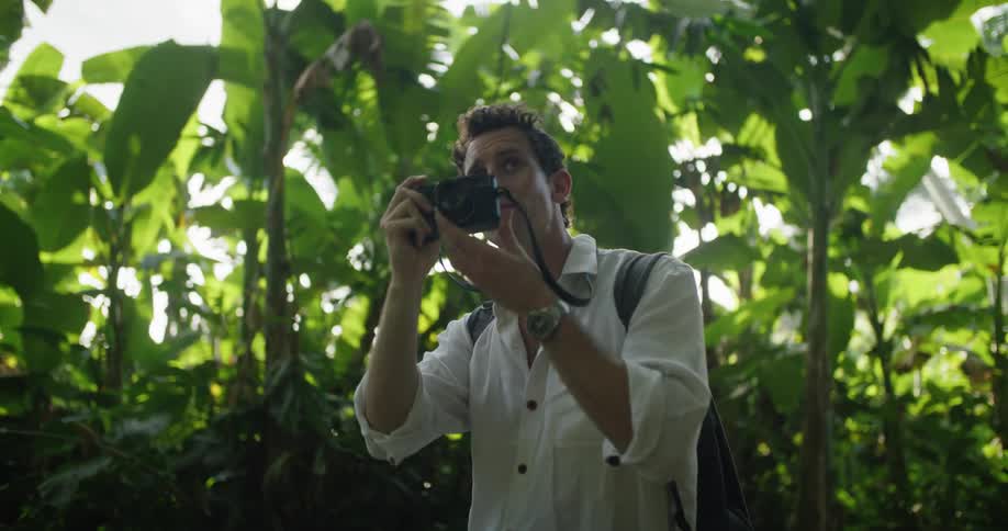 A man is standing in a tropical jungle and he lifts his camera to take a picture as the sun shines brightly behind him