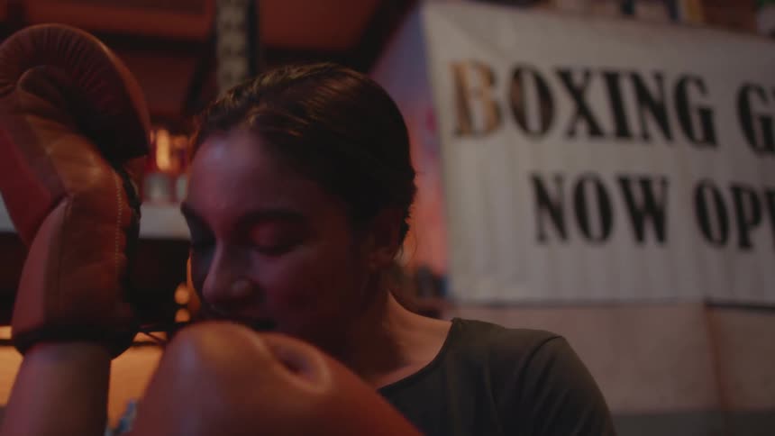 A woman uses her teeth to pull and help tighten the laces on her boxing gloves in the gym
