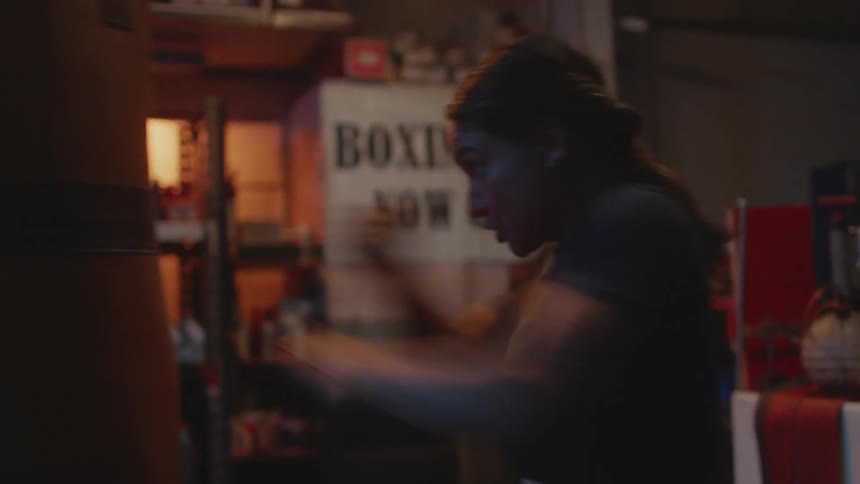 A woman circles a punching bag while striking it with her fists in a dimly lit boxing gym