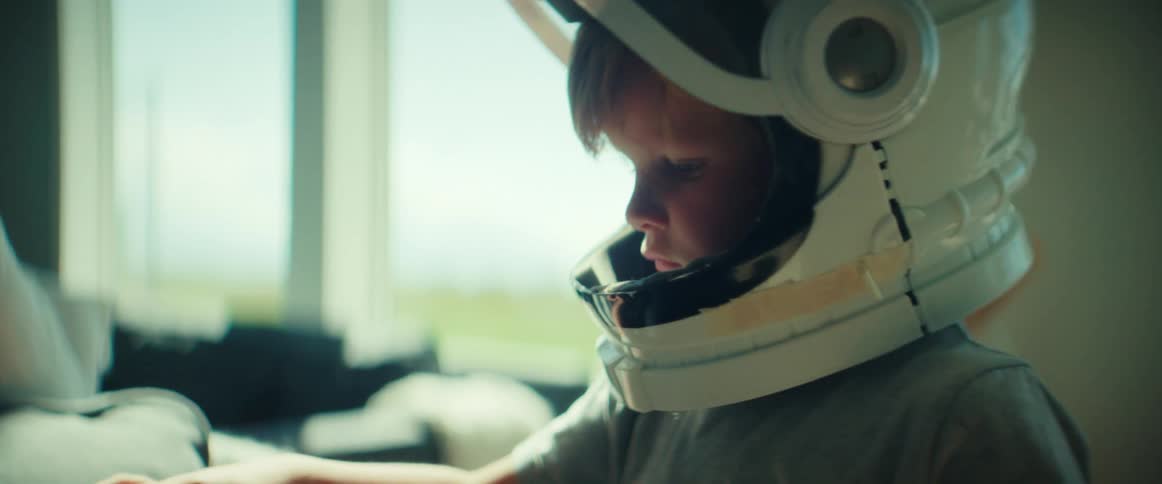 A boy wearing a toy astronaut helmet plays with a toy car in the living room of a house during the day
