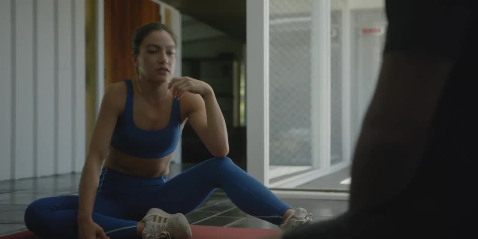 A woman in exercise clothing sits on a yoga mat and talks with someone
