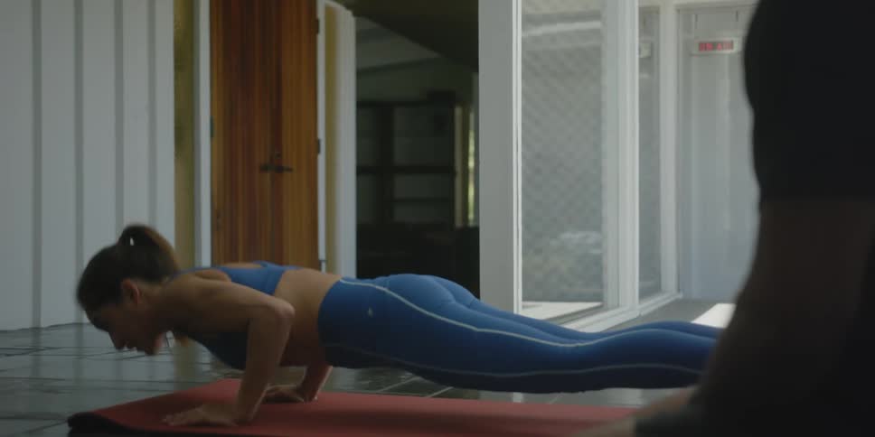 A woman does exercises on an exercise mat while talking to a man sitting on the floor nearby
