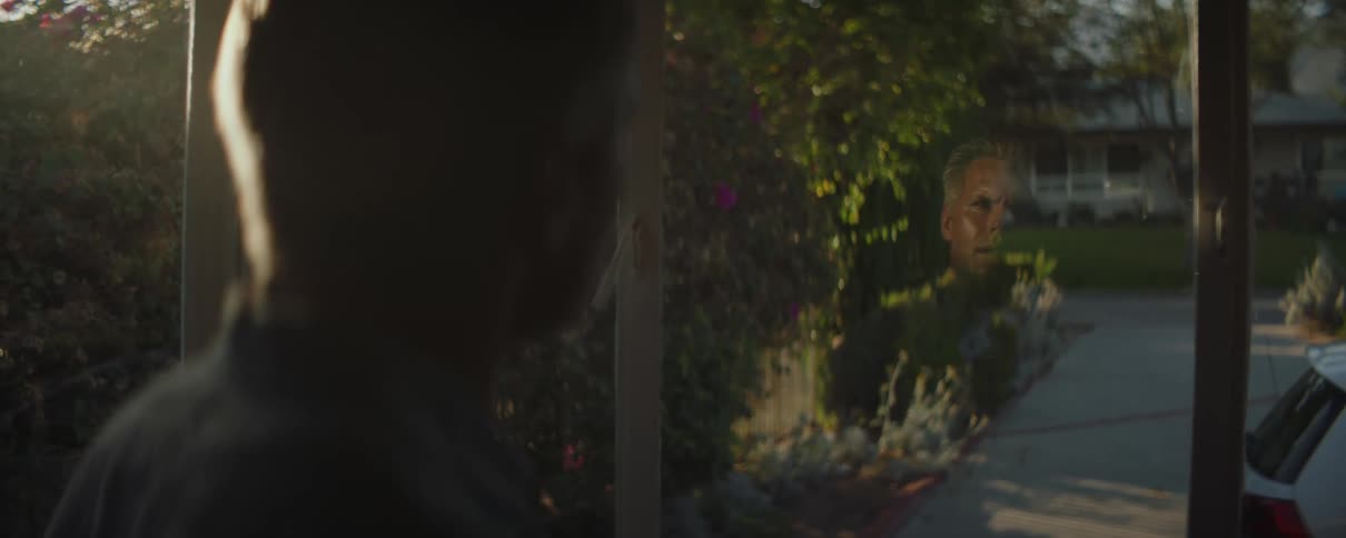 A man is standing inside looking at his reflection in a window facing a driveway
