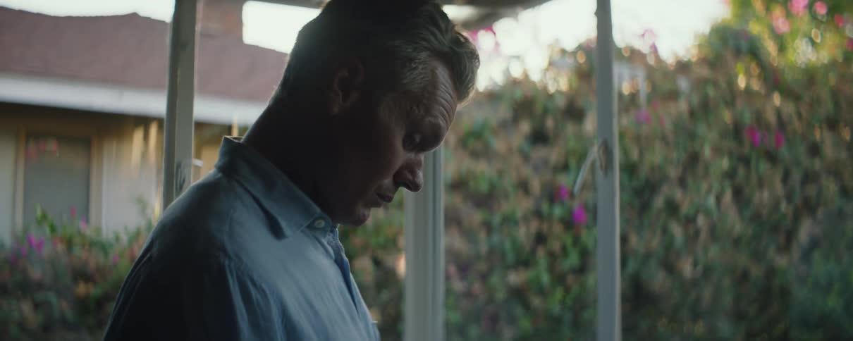 Man with blue shirt looks down and smiles slightly in living room next to windows overlooking street