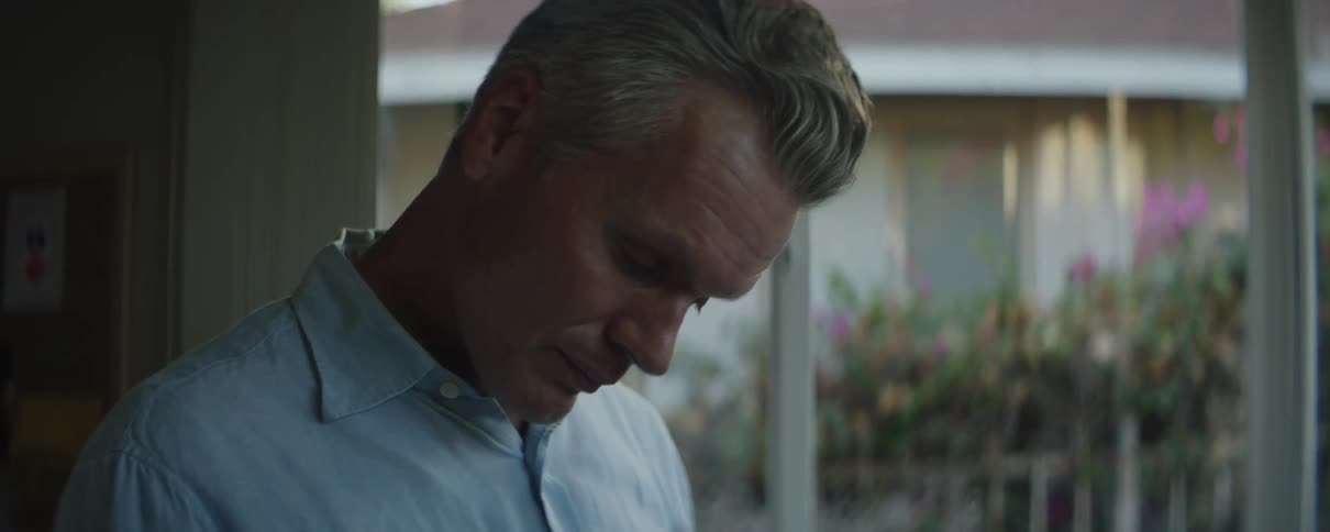 A man is standing in a room near windows looking down and smiles then rubs his nose with his hand