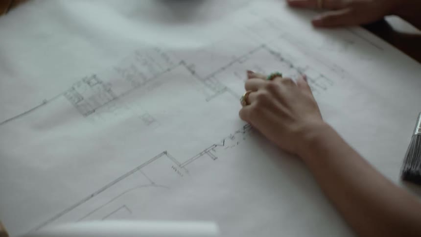 Drafting plans are on a table as a woman moves her hand pointing to an area a man places his hand on top of hers and holds her hand