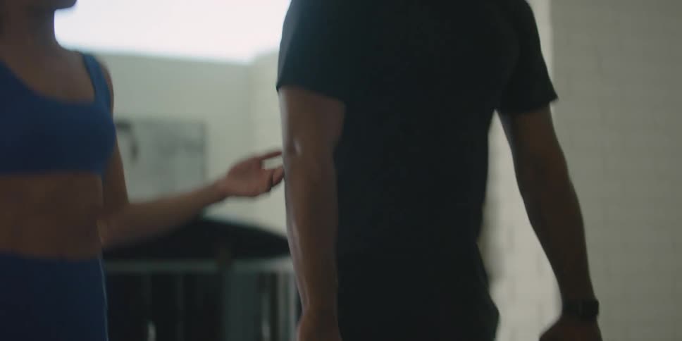 Man with shaved head glasses beard and black shirt lifts barbell next to woman with pony tail and blue top in the gym
