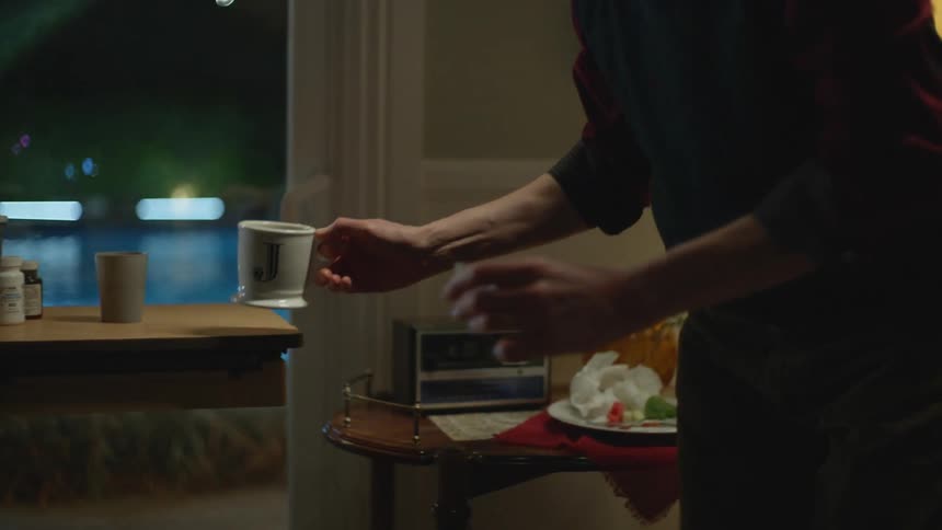 A man at home picks up a beverage and some pills and passes it to an ill person in a bed hooked up to medical devices
