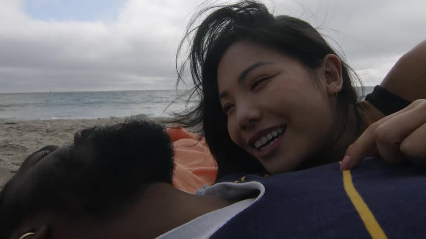 A man and woman lie on a beach and talk as she rubs her hand on his chest