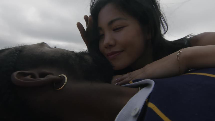 A man and woman lie on a beach and she rests her hand on his chest as he rubs her hair off her face