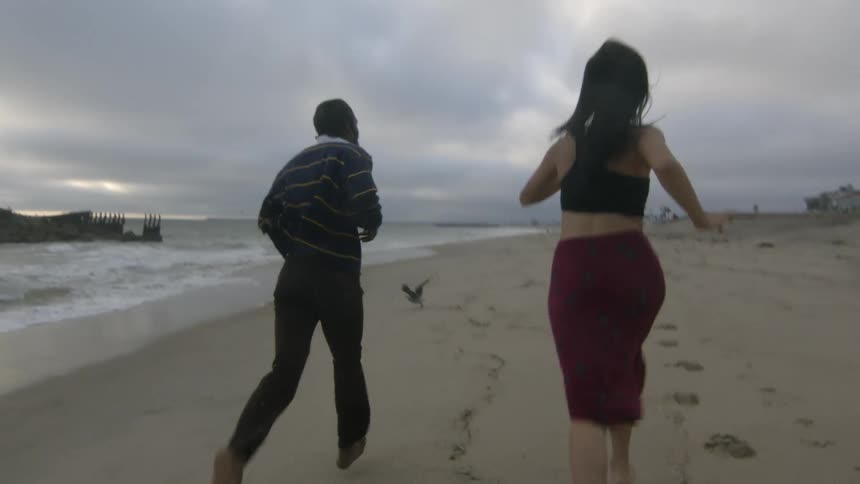 Man with stripped sweater black pants and woman wearing black top and red skirt runs along the beach next to the ocean