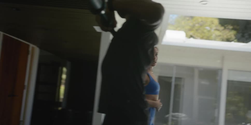 A man lifts weights and a woman wearing workout gear walks past him and talks
