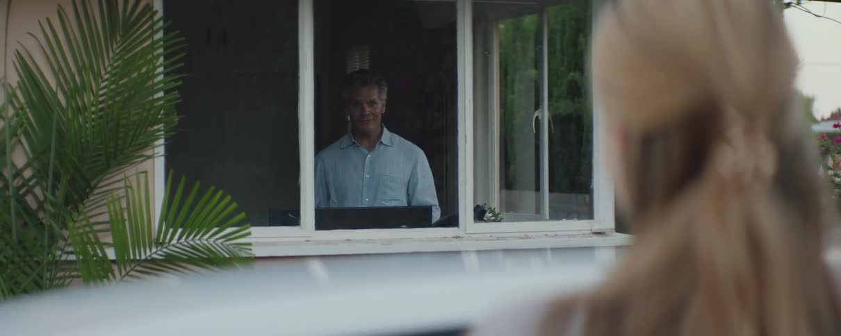 A man is inside the house looking out the window and when he sees his teenage daughter looking at him from outside he smiles
