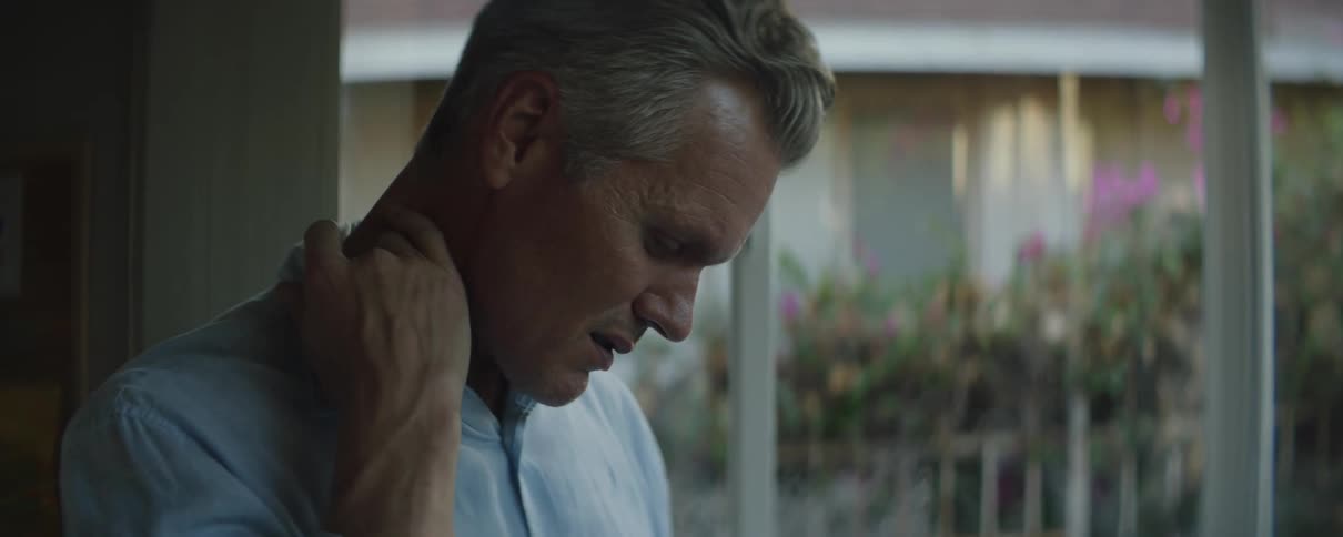 Older man stands near windows looking down with a frown
