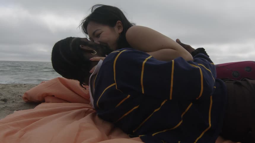 A man and woman share a kiss while lying on a blanket on the beach on a cloudy evening