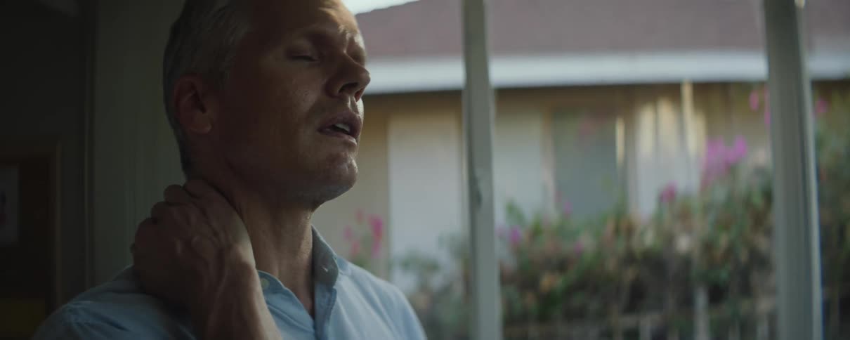 A man rubs his neck while standing next to a window