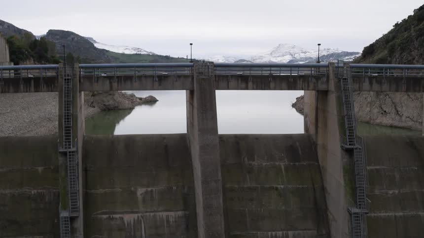 Water and mountains are seen beyond a tall dam just before the camera pans down the dam wall