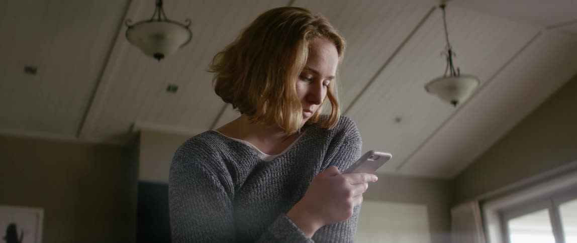 Young woman wearing a comfortable sweater stands on the living room at home looking at her phone