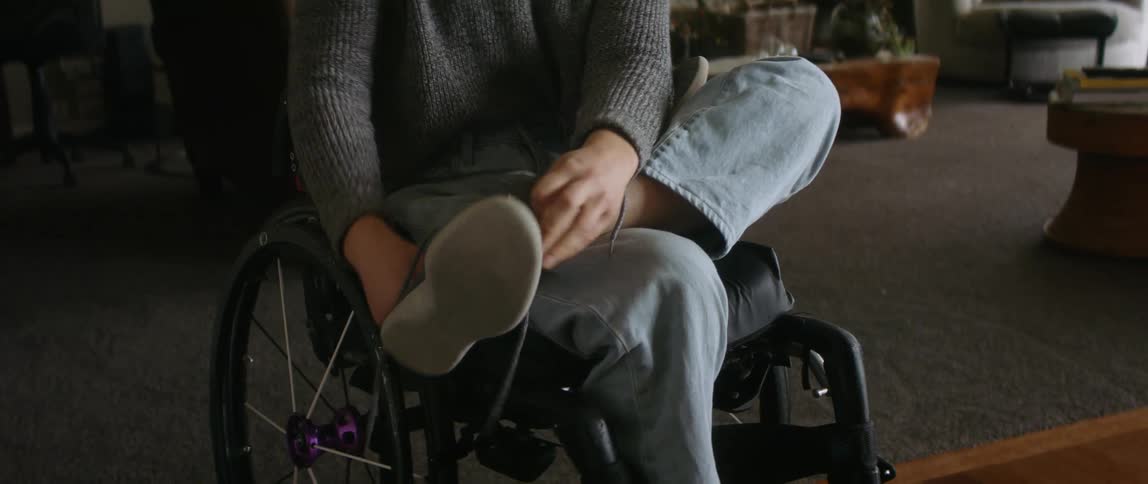 Blonde woman sitting in wheelchair in the living room puts on a shoe