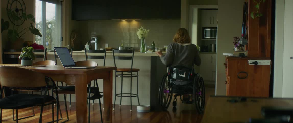 A girl in a wheelchair is sitting at the kitchen table with a laptop computer and takes her coffee cup and rolls around the counter