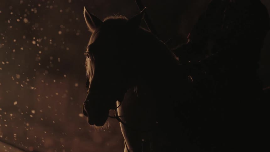 A man holding a sword rides a horse past a fire at night