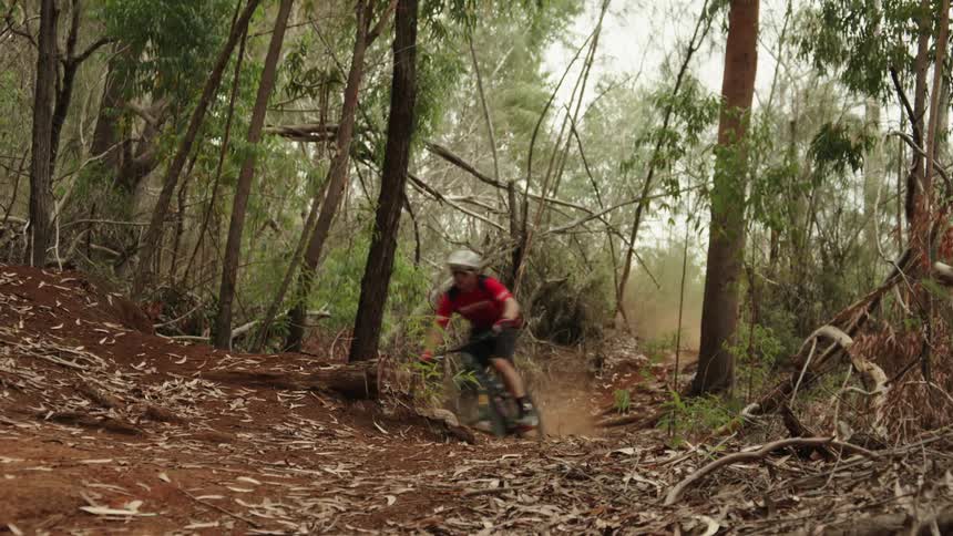 A cyclist goes airborne and does a trick while speeding down a dirt trail in a woodland during the day