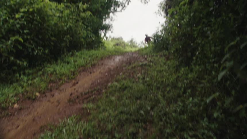 A cyclist does a bicycle trick in the air while riding along a dirt trail at high speed on a rainy day