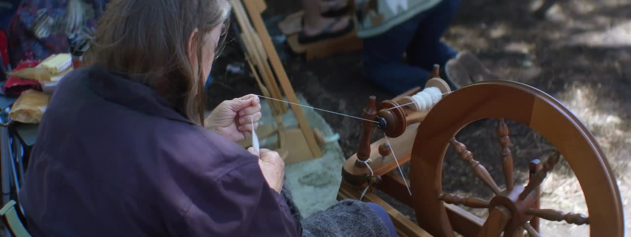A woman feeds thread into a spinning wheel while sitting outdoors during the day