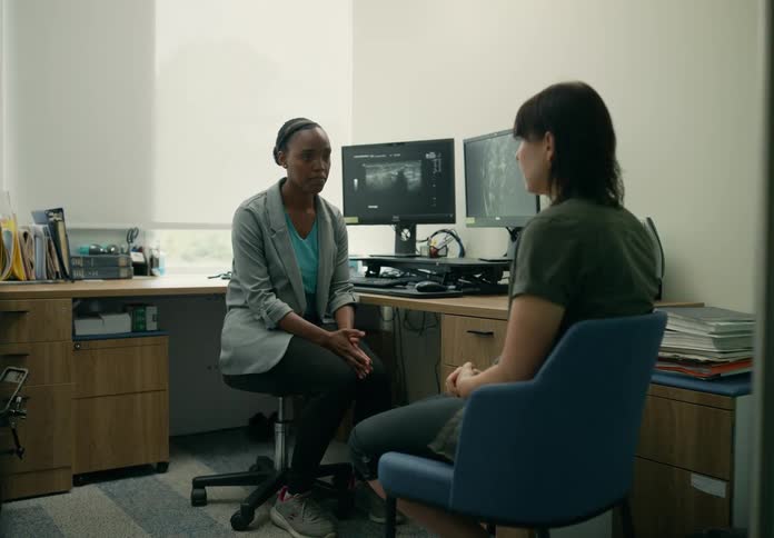 A woman sits in an office with medical results on computer screens as a doctor discusses them with her