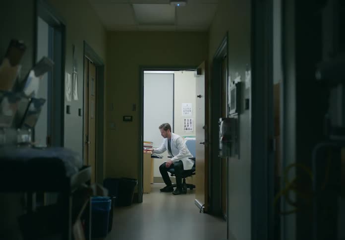 A doctor yawns while typing on a computer in an examination room before turning off the light and leaving