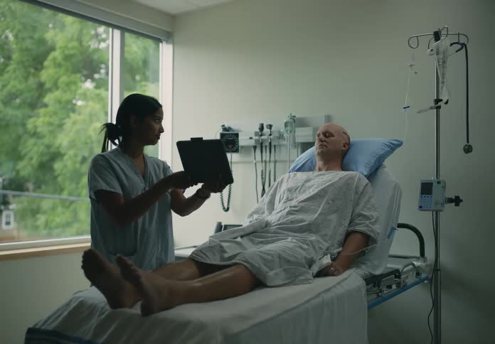 Man with scar on his head lying in bed in the hospital while a nurse is standing next to him holding a tablet for him