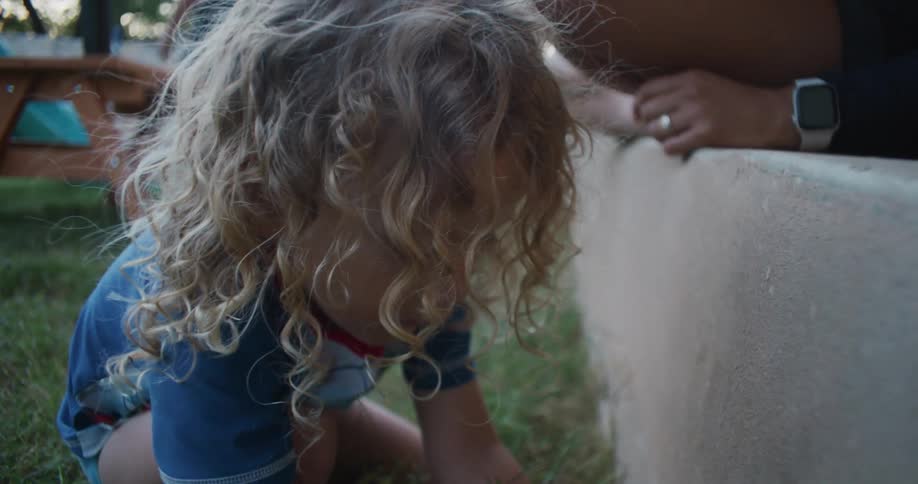 Blonde girl plays in grass as another person sits on low wall nearby