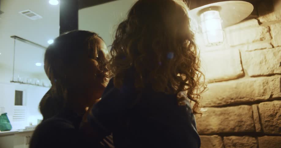 Mother with ponytail holds daughter with curly hair in her arms next to halogen lamp