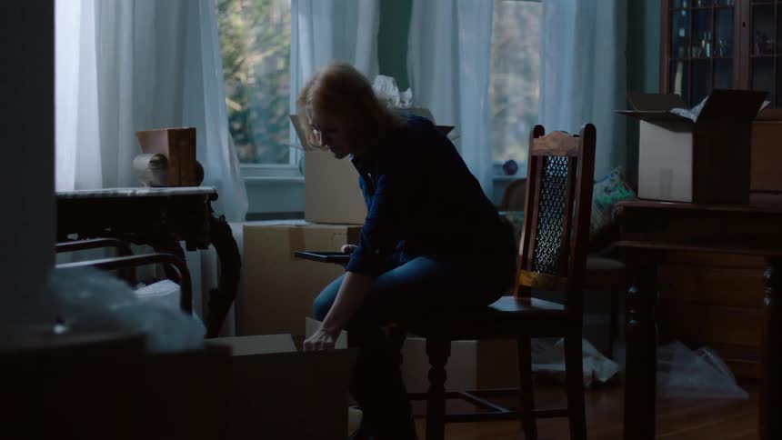A woman walks to a table and picks up a picture in a frame and sits down in a chair next to a stack of boxes and wraps the picture and puts it in a box