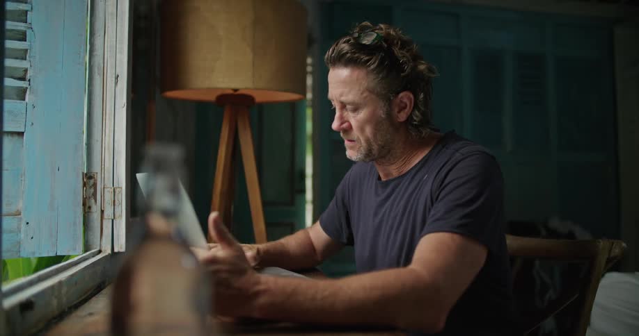A man is sitting at a table in a room typing on a laptop computer in front of an open window