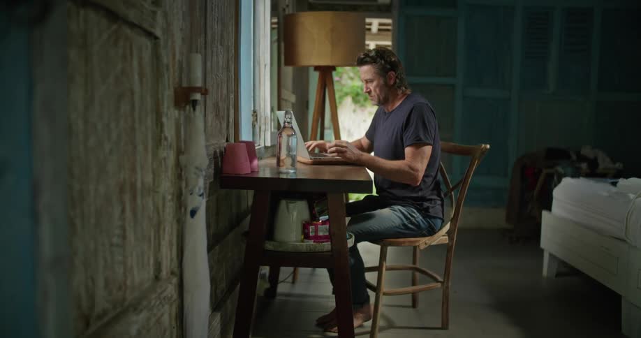A man is sitting at a table in a room typing on a laptop computer in front of a window