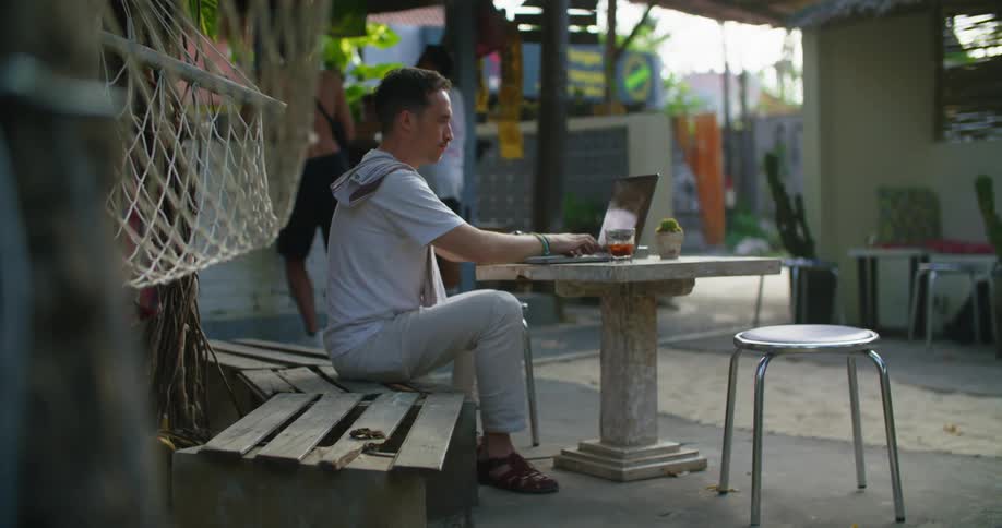 A man is sitting at a table at an outdoor cafe typing on his laptop computer and drinking from his glass as traffic moves on the street