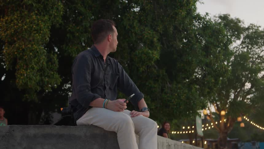 A man is sitting on a concrete slab outside doing something on his phone that causes him to look satisfied