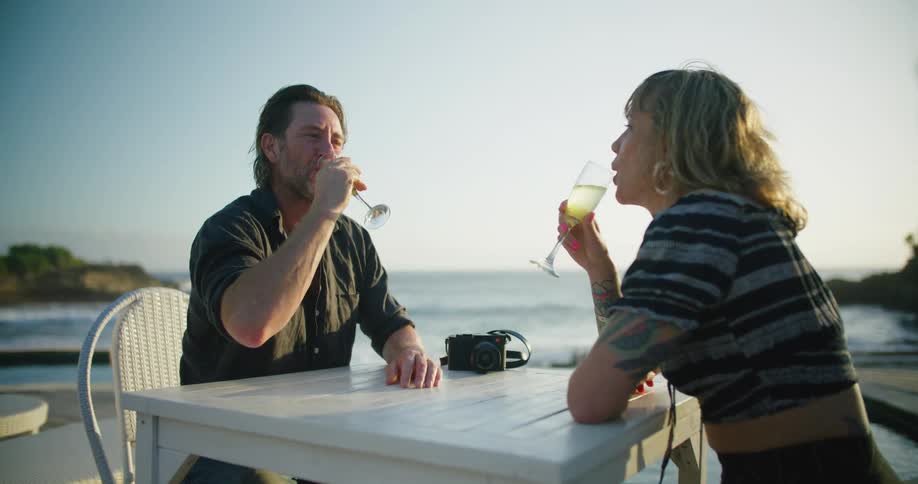 A couple toast and drink champagne while sitting outside at a seaside restaurant